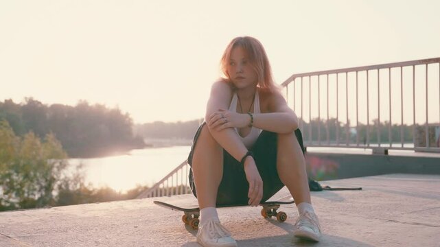 Bold Skater's Twilight Pose: Redhead Teen Girl Frontal on Board | Backlit Urban Scene by Sunlit River | Slow Motion Portrait on Skateboard with Daring Attitude and Bridge Stairs