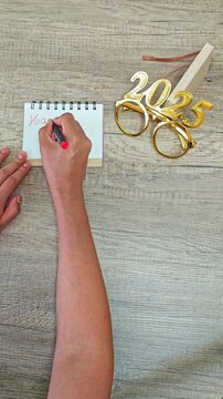 Woman hand writing happy new year 2025 with red pen on notebook paper on wooden desk. Next to golden new year 2025 glasses.