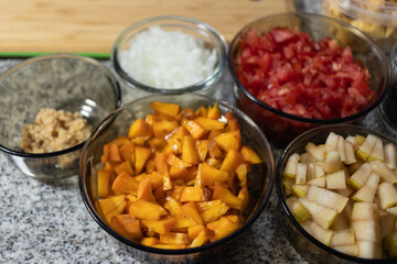 Ingredients to prepare chiles en nogada. Various chopped vegetables, fruits, mango and seeds in glass bowls on a gray countertop next to a wooden cutting board.