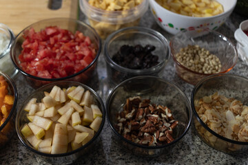 Ingredients to prepare chiles en nogada. Various chopped vegetables, fruits, nuts, and seeds in glass bowls on a gray countertop next to a wooden cutting board.