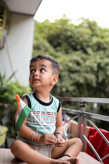 Cute Indian boy holding Indian National Flag in his hangs at home balcony. Bright portrait of happy child sitting on the table with national flag