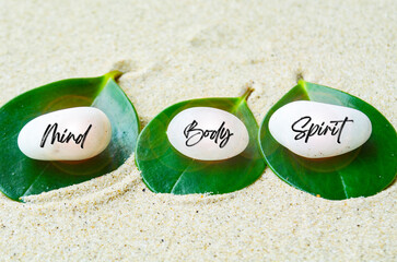 Mind, Body and Spirit text engraved on white zen stones with leaf and sand background. Mind, Body...