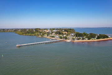 Fototapeta premium Aerial View of a Coastal Pier Extending into Calm Waters, Small Town Shoreline and Blue Sky Background at Wellington Point, Queensland, Australia