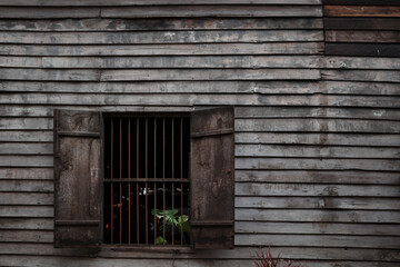 A close-up of a weathered wooden wall featuring an open shuttered window with metal bars. 