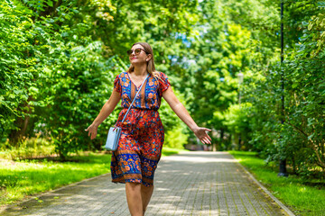 Beautiful blonde woman wearing colorful floral flowing dress walking with hands spread out on green background in city park on sunny day. Front view