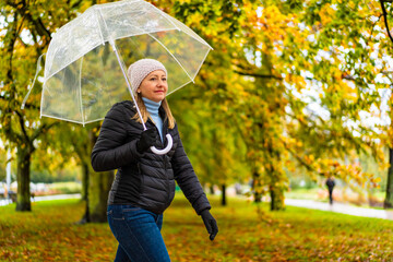 Middle aged woman wearing black jacket, jeans and cap walking with transparent umbrella in city park on autumn rainy cool day. Side view