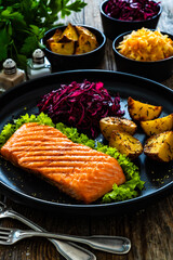 Grilled salmon steak with baked potatoes and red cabbage on black plate on wooden table
