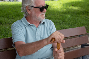 Smiling white haired elder 70s man with walking disability holding knob of cane,sitting  in a bench...