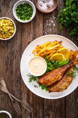 Fried sea bass served on lettuce with French fries, lemon and mayonnaise on white plate on wooden table
