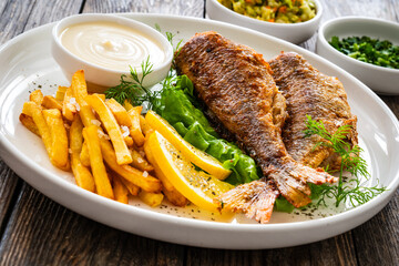 Fried sea bass served on lettuce with French fries, lemon and mayonnaise on white plate on wooden table
