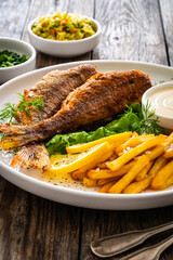 Fried sea bass served on lettuce with French fries, lemon and mayonnaise on white plate on wooden table
