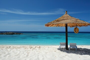 A thatched umbrella provides shade on a pristine white sand beach with two lounge chairs and a straw hat hanging from the umbrella. 