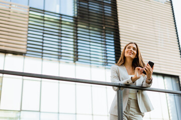Fototapeta premium Smiling business woman enjoying a sunny day outdoors while texting on her smartphone near modern architecture in an urban setting