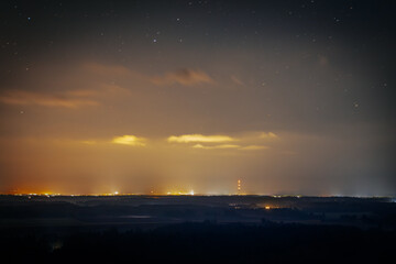 A serene night sky filled with stars stretches over a distant horizon illuminated by the soft glow of city lights. The contrast between the dark landscape and the warm light creates a peaceful