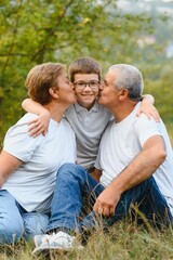Obraz premium Grandson, grandfather and grandmother sitting on grass having fun outdoors in park at sunset