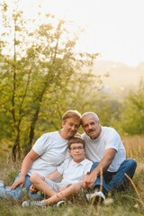 Fototapeta premium Grandson, grandfather and grandmother sitting on grass having fun outdoors in park at sunset