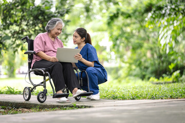 Elderly woman in a wheelchair and a nurse sharing a moment, exploring something on a laptop in a peaceful park setting