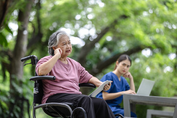 Obraz premium Elderly woman in a wheelchair expressing concern during a conversation, while a nurse looks on, in a serene outdoor setting.