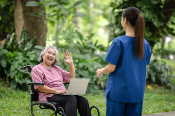 Elderly woman in a wheelchair waving with a smile while holding a laptop, engaging warmly with a nurse in a green park.