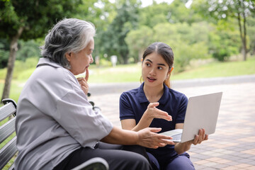 Fototapeta na wymiar Young caregiver discussing senior care plans with an elderly woman using a laptop while sitting on a park bench, emphasizing attentive support and personalized elderly care in a serene environment.