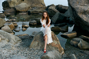 Serene woman in flowing white dress sitting with legs crossed on rocky shore by peaceful water