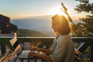Young woman sitting on balcony with view of mountains and sunset working using laptop