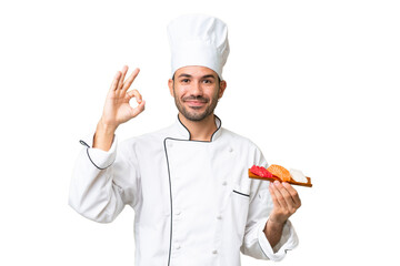 Young caucasian chef holding a sushi over isolated background showing ok sign with fingers © luismolinero