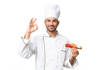 Young caucasian chef holding a sushi over isolated background showing ok sign with fingers