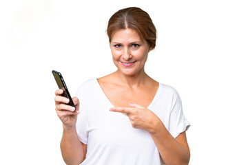 Fototapeta premium Middle-aged caucasian woman over isolated background using mobile phone and pointing it