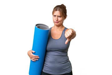 Middle-aged sport woman going to yoga classes while holding a mat over isolated background showing thumb down with negative expression