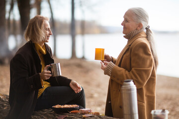 Two woman friends  drinks tea and enjoying a holiday  in autumn outdoor.  portrait of a smiling senior woman with an adult daughter relaxed outdoors 