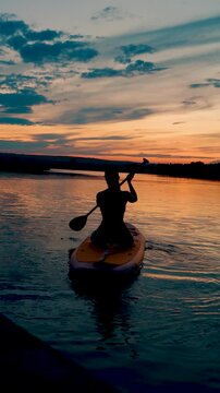  A young woman swims on a SUP board in a lake at sunset. Silhouette of a woman sitting on a SUP board at sunset. SUP surfing concept.Vertical video
