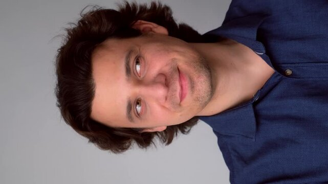 Portrait of man looking by sides with sly tricky face on gray background. Curly middle aged guy having tricky plan in mind with smile. Wearing blue shirt. Showing emotions through facial expressions.