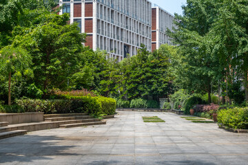 A modern, clean, and contemporary urban park featuring an inviting outdoor space. A paved walkway, lined with lush green hedges and trees, a staircase with a sleek, modern building in background