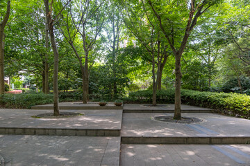 Urban park with a modern design with elevated stone platforms, creating a terraced effect.Green trees and shrubs providing shade and a natural atmosphere.Peaceful and inviting outdoor space