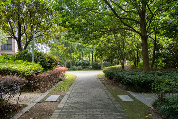 A paved pedestrian walkway winds through a lush public garden, surrounded by landscaped shrubs and trees that provide shade. This urban parkland within a residential community of apartment blocks