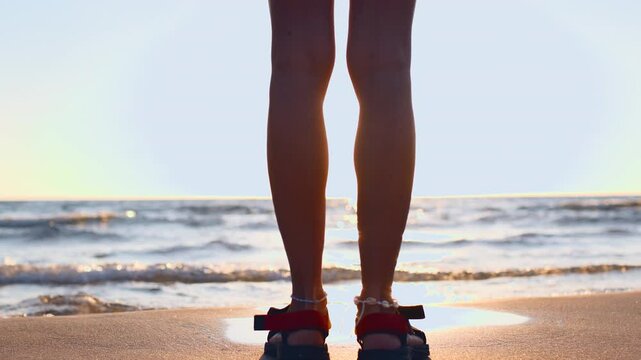 slender girl in sandals stands on the sand and looks at the sea.  summer mood. girl's legs close-up. girl on the beach.