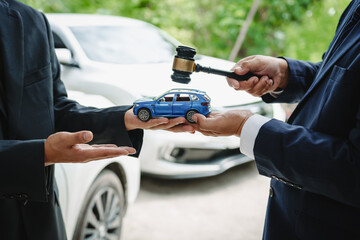 Two Asian individuals discuss a car insurance claim with an agent in front of a damaged vehicle. They review documents, discuss the policy, and address the crash and repair costs.