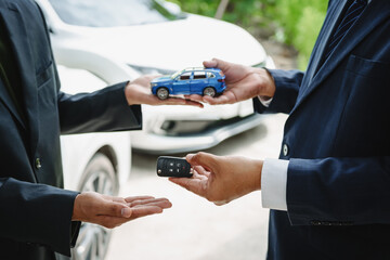 Two Asian individuals discuss a car insurance claim with an agent in front of a damaged vehicle. They review documents, discuss the policy, and address the crash and repair costs.
