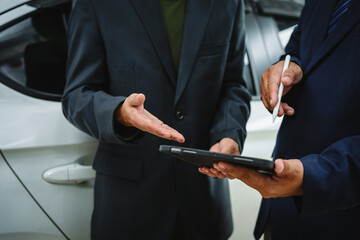 Two Asian individuals discuss a car insurance claim with an agent in front of a damaged vehicle. They review documents, discuss the policy, and address the crash and repair costs.