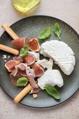 Olive-colored plate with ricotta cheese, figs, prosciutto, grissini and fresh basil, vertical shot, middle close-up, elevated view