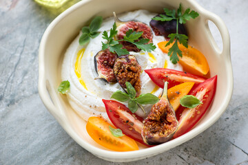 Ricotta dipping sauce with torn figs and tomato wedges in a beige serving tray, horizontal shot on a grey granite surface