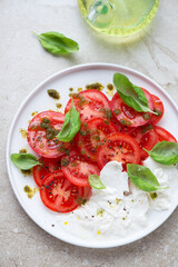 Red tomatoes with basil oil, fresh basil and burrata cheese, vertical shot on a beige stone surface, high angle view