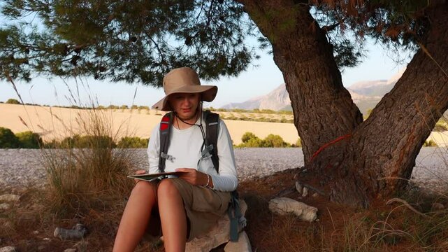 Hiker sits under tree, consulting map while enjoys hade. Girl Wearing hat and backpack,  hiker takes moment to plan  next adventure in serene outdoor setting