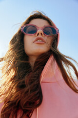 Stylish woman in pink hoodie and sunglasses posing against vibrant blue sky
