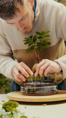 Side-view of young male preparing moss for a glass terrarium at home workshop with plants in the background. Home gardening, greenery business concept
