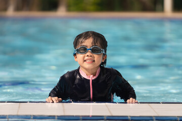 A young girl is in a pool wearing a black swimsuit and goggles. She is smiling and looking at the camera