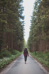 Fototapeta premium Young woman in a red plaid shirt walking down a peaceful, tree-lined forest road, embracing the tranquility and beauty of nature, symbolizing freedom and a journey of self-discovery