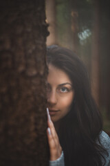 Fototapeta premium Enigmatic woman with captivating blue eyes peeks from behind a tree in a mystical forest. The image captures the essence of mystery, nature, and serene beauty