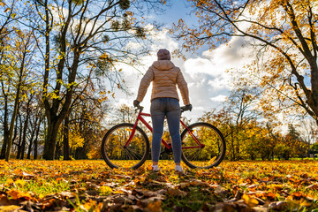 Naklejka premium Woman wearing beige jacket, jeans and winter hat standing with red bicycle on clearing in forest in autumnal scenery on sunny day. Back view